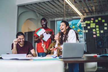 Modern yaratıcı insanlar birlikte çalışırken çılgınca komik fikirlere gülüyorlar ve kağıtlar ve dizüstü bilgisayarlarla masaya oturuyorlar ve iş projesinin sunumuna hazırlanıyorlar.