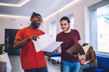 Günlük giysiler ve gözlüklerle erkeklere taslak gösteren odaklanmış siyahi bir adam. Not defteri paylaşım stratejisi ile ofis alanında yürüme stratejisi.
