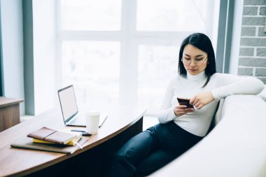 Uzun saçlı, gözlüklü, beyaz balıkçı yakalı, cep telefonlu akıllı Asyalı kadın. Masadaki koltukta dizüstü bilgisayarla oturuyor. 