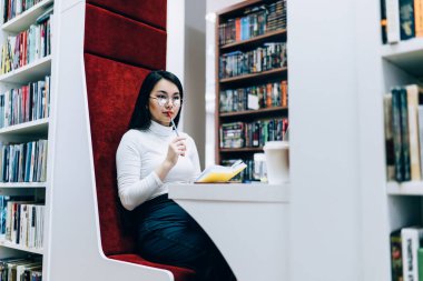 Kitap okurken düşünen ve dizüstü bilgisayarla kütüphanede rahat bir şekilde oturan sakin Asyalı kadının yan görüntüsü