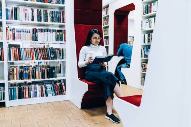 Sıradan giyinmiş konsantre Asyalı kadın ve modern kitapçıda rahat kırmızı koltuklarda oturan ve kitap okuyan yüzü olmayan adam. 