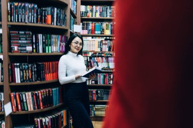 Genç etnik esmer, gözlüklü, elinde açık kitaplıkla kameraya bakıp gülümserken kitap rafına yaslanmış. 