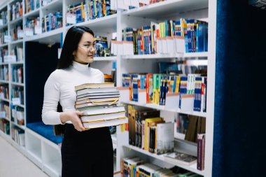 Siyah saçlı, beyaz bluzlu, gözlüklü genç Asyalı bir kadın kütüphanedeki sıra sıra kitap raflarının arasına kitap yığını taşıyor.