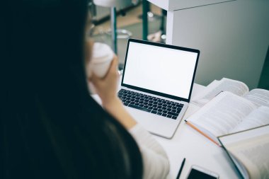 Beyaz ekran ve kitaplarla çalışırken masada oturup kahve içen siyah saçlı kadının arka plan görüntüsü.