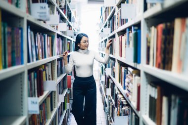 Asyalı genç bir kadının seçici odak noktası beyaz yaka gömlek ve siyah pantolon giymesi kütüphanede kitaplıkların arasında dolaşması ve kitap araması.