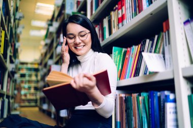 Koyu renk saçlı Asyalı kadın bir eliyle yuvarlak bardaklara dokunuyor, diğer eliyle kitabı açıyor ve kütüphanede kitaplıklar arasında oturuyor.