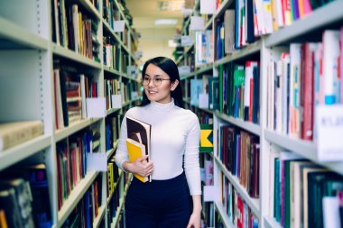 Siyah saçlı, mutlu, yuvarlak gözlüklü, renkli defterleri olan Asyalı kız öğrenci kitaplıkların arasında yürüyor ve kütüphanede kitap arıyor.