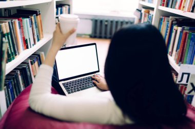 Siyah saçlı kadının arka planda oturduğu koltukta elinde sıcak içecekle kağıt bardakla boş ekranlı dizüstü bilgisayar kullandığı sahne.