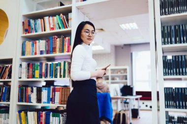 Cep telefonu olan düşünceli, uzun saçlı Asyalı bir kadının kütüphanede kitaplarla dolu raflarda yürümesi.