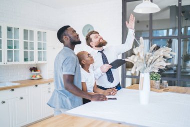 İlgilenen Afro-Amerikan çiftin resmi kıyafetli ve kendine güvenen bir adamın geniş modern bir evin mutfağında durmuş tavanı işaret edişinin yan görüntüsü.