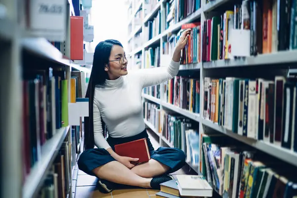 Tebessüm eden Asyalı genç kadın kütüphanede ders arıyor ve çapraz bacaklarla yerde otururken kitaplıktan kitap topluyor.