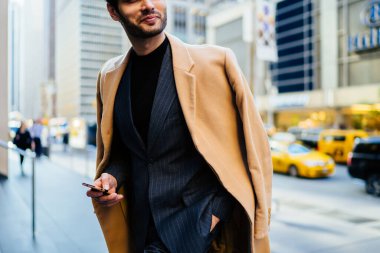 Deneyimli erkek girişimci zarif giyinmiş metropoliten şehir ortamında duruyor, resmi giyinmiş başarılı bir iş adamının modern akıllı telefon teknolojisini elinde tutarken görüntüsü.