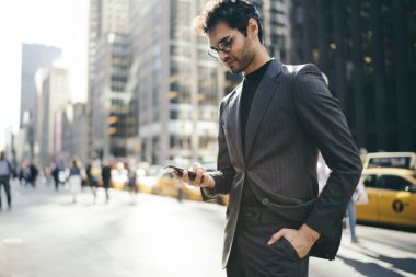 Resmi giysiler içinde Avrupalı erkek girişimcinin şehir merkezinde ayakta duran akıllı telefondan SMS mesajı yazışının yan görüntüsü, mobil uygulamalara bağlı olarak finansal haberler okuyan başarılı ekonomist