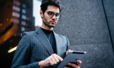 Başarılı erkek girişimci zarif giysiler içinde modern dokunmatik ped cihazıyla ilgili haberleri indirirken 4G internet bağlanıyor ofis binasının yanında duruyor, tarama yapıyor.