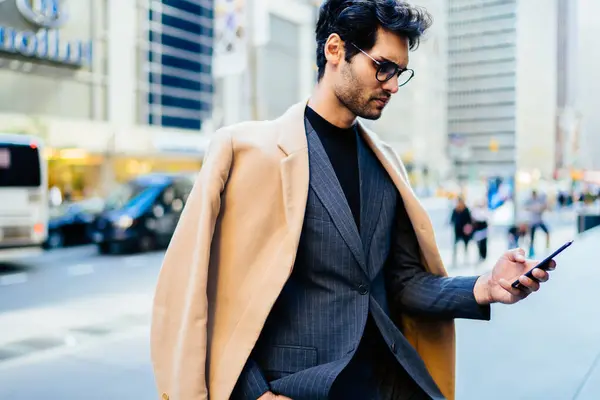 Confident businessman in elegant outfit reading income message with good news on smartphone standing on street, male entrepreneur in optical glasses checking tariffs for roaming internet on mobile