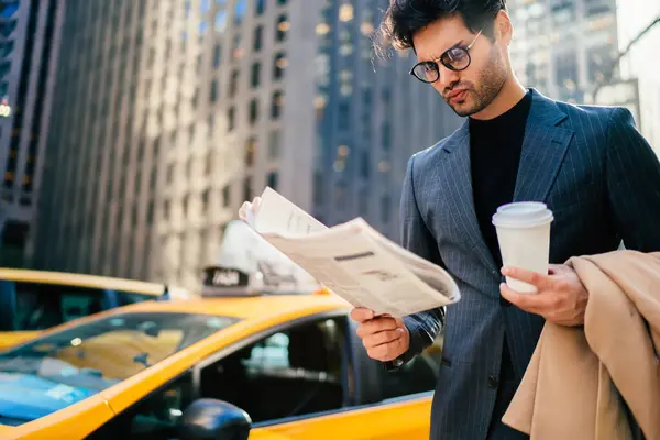 Puzzled male economist searching article with financial news in morning newspaper holding tasty coffee to go in hand, pensive manager in formal wear reading business press standing at urban setting