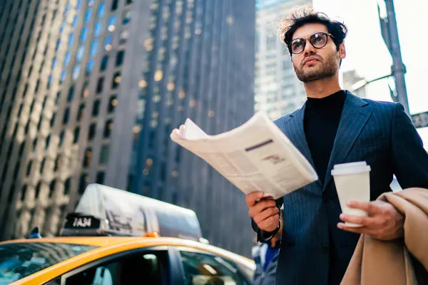 Below view of puzzled male employer in classic spectacles thinking on information from newspaper article standing at urbanity in downtown, serious businessman pondering on journal fresh news