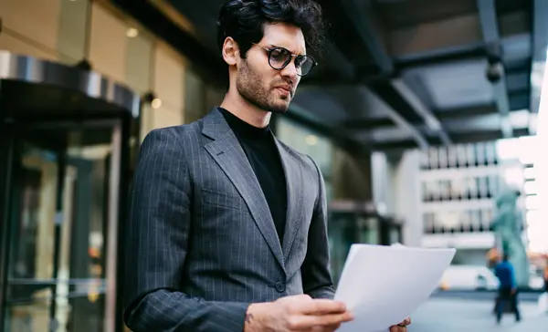 Puzzled lawyer dressed in stylish formal apparel reading financial reports standing near office building, concentrated proud CEO holding corporate documents and analyzing productivity for strategy