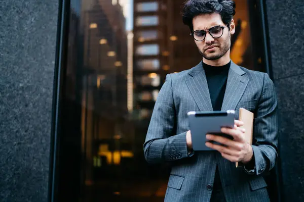 Puzzled male lawyer in formal wear typing text on digital tablet device to publicate information on corporate internet website standing outdoors, businessman solving problem with web banking