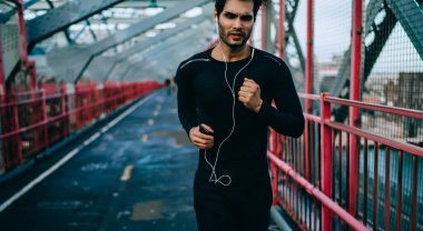 Çevrimiçi web podcast 'ten müzik motivasyonu çalma listesinin keyfini çıkaran aktif sporcu, elektronik kulaklıkla ses kayıtlarını dinlerken şehir köprüsünde koşan genç erkek sporcu