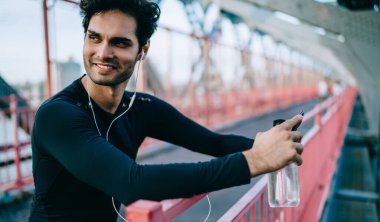 Elektronik kulaklıklı neşeli erkek koşucu şehirde sabah antrenmanının keyfini çıkarıyor. Kenar mahallede duruyor ve kulaklıklı mutlu adam pozitif müzik podcast 'i dinliyor ve dışarıda gülümsüyor.