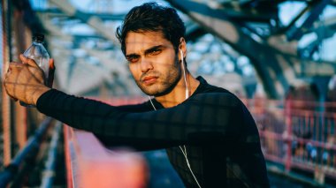 Avrupa 'nın atletik vücut geliştiricisinin portresi kentte duruyor ve kameraya bakıyor refah eğlencesi ve sağlıklı yaşam tarzı hakkında sesli kitap dinliyor, elektronik kulaklık takan genç bir koşucu