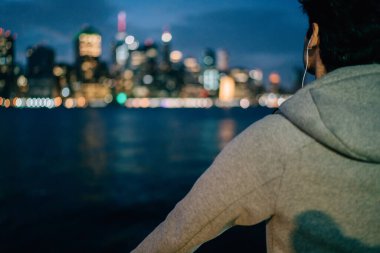 Hudson Nehri 'nin kıyısında modern kulaklıklarla New York şehir manzarasında bulanık manzarası olan ve sesli kitap dinleyen düşünceli bir erkek müzik sever.