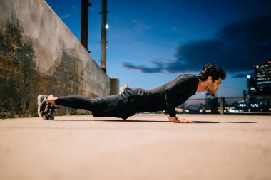 Kenar görüşlü kaslı sporcunun akşam saatlerinde şehirde yaptığı itme egzersizleri elektronik kulaklıkla motivasyon ses kitabı dinlemesi, erkek sporcunun açık havada kardiyo sporuyla oynaması.