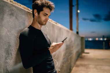 Eşofman giymiş, modern kulaklıklarla müzik dinleyen sağlıklı bir adamın yan görüntüsü. Akşamları şehir dışında antreman yapan, cep telefonuyla sohbet eden Avrupalı erkek vücut geliştirici.