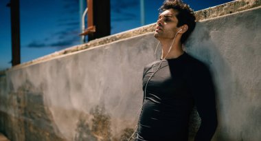 Eşofman giymiş, kulaklıkla müzik dinleyen yorgun, gece antrenmanından sonra duvara yaslanan, açık havada spor yaptıktan sonra etkin bir şekilde koşan yorgun bir adam.