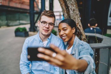 Neşeli Afro-Amerikalı kadın, beyaz tenli hipster adamla birlikte kameranın önünde poz veriyor. İnternet blogu için fotoğraf çekiyor. Şehir ortamında oturuyor, çeşitli arkadaşlar selfie çekiyor.