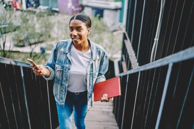 Koyu tenli, sıradan giyinmiş hippi kız cep telefonuyla mesajlaşırken kameraya bakıyor. 4G internet kullanıyor. Cep telefonuyla merdivenlere tırmanan mutlu Afro-Amerikalı genç bir kadının portresi.