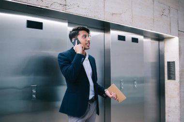 Düşünceli Hindu erkek gururlu akıllı ceo cep telefonuyla konuşuyor içeride asansörlerin yanında duruyor, kendine güveni tam, resmi kıyafetli iş adamı servis destek operatörüne akıllı telefon görüşmesi yapıyor.