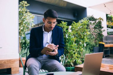 Yakışıklı, şık giyimli ve gözlüklü erkek girişimci. Kafe terasında uzaktan çalışırken notlar alıyor. Kendine güveni tam, zengin bir blog yazarı.