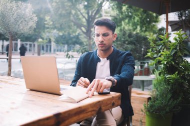 Moda kıyafetli ve gözlüklü kendine güvenen Hintli erkek blogcu kafeterya terasında blog yayını yapma fikri üzerinde kafa yoruyor. Ciddi iş adamı, uzakta çalışırken dizüstü bilgisayarını kullanıyor.