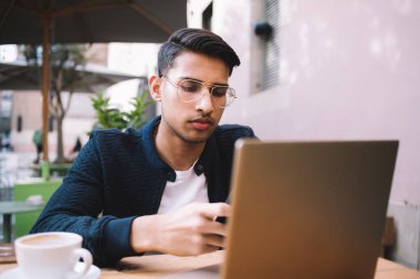 Moda gözlüklü ciddi yakışıklı Hindu erkek. Uzaktan kumandalı bilgisayar ve kablosuz bağlantı kullanıyor. Düşünceli iş adamı banka hesaplarını ve işlemlerini cihazlarla kontrol ediyor.