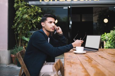 Ciddi müreffeh Hindu milenyum erkek serbest çalışan cep telefonuyla iş konuşan yetenekli bir adam bilgisayarla kafeterya terasında çalışıyor ekran araması yapıyor.