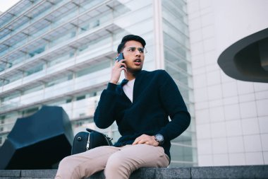 20 'li yaşlarda şık giyinmiş, şık giyinmiş, şehir merkezinde meydanda oturmuş, akıllı telefon görüşmesi yapan, gezgin, yakışıklı, hippi erkek gezgin, şehir merkezinde cep telefonuyla konuşuyor.