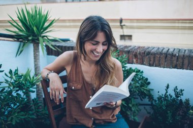 Yukarıdaki pozitif kadın, günlük kıyafetlerini giymiş kitap okuyor ve terasta, yeşil saksı bitkilerinin arasında tahta sandalyede oturmuş gülüyor.