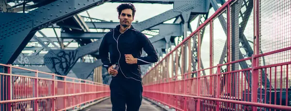 Active sportsman running with bottle of water in hand and enjoying music motivation playlist, young male athlete jogging on city bridge during listening audio records via electronic headphones