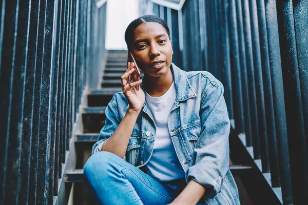 Portrait of attractive female making international cellphone conversation during free time at urban setting, youthful hipster girl with dark skin looking at camera while communicating via smartphone