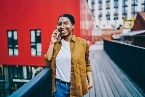 Smiling dark skinned hipster girl in spectacles satisfied with tariffs for mobile calls in roaming talking on cellphone, portrait of cheerful African American woman making conversation on smartphone