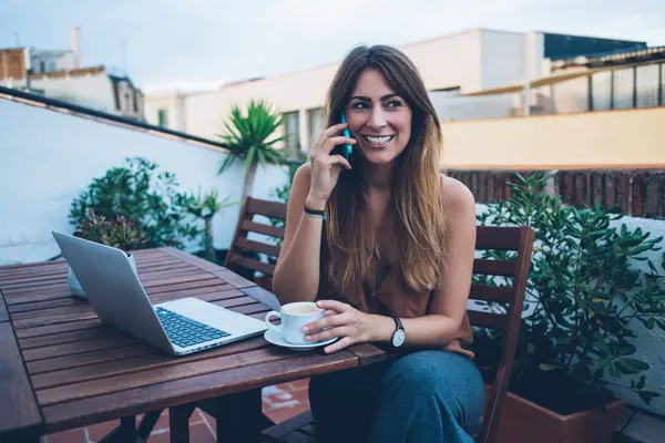 Mutlu serbest çalışan bir kadın bilgisayar ve cappuccino ile masada oturuyor ve telefonda konuşurken gülümsüyor ve başka tarafa bakıyor.