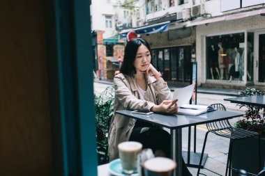 Meraklı Asyalı kadın elinde kağıtlarla çalışıyor ve elinde açık bir defterle caddedeki kafede oturuyor. 