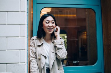 Neşeli gülümseyen, sıradan, trençkotlu Asyalı bir kadın telefonda konuşurken güpegündüz sokak girişinde duruyor.