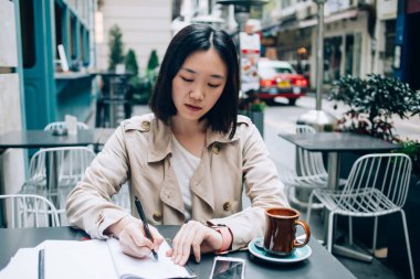 Özenli, kısa saçlı, bej yağmurluk giyen Asyalı bir kız öğrenci sokak kafesinde oturmuş ödev yapıyor deftere yazıp kahve içiyor.