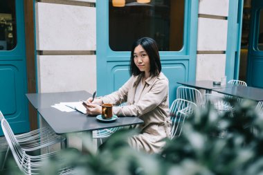 Kısa saçlı, bej yağmurluk giyen Asyalı bir kız öğrencinin sokak kafesinde oturup telefonla mesajlaşarak kameraya bakışının yan görüntüsü.