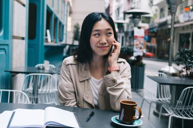 Genç Asyalı içerikli kadın cep telefonuyla konuşuyor ve gülümsüyor kahve ve not defteriyle kentin arka planında dinlenirken.