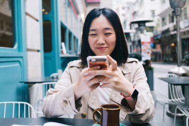 Neşeli, güzel, modern, ceketli, akıllı sörf arabalı bir Asyalı kadın. Masada oturmuş bulanık caddenin arka planında çay içerken. 