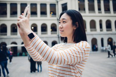 Modern akıllı telefona bakan ve gezideyken şehir caddesinin fotoğrafını çeken genç bir etnik kadının yan görüntüsü